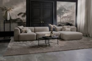 Element sofa Emanuelle with a longchair and an ottoman between the elements. Made from a light beige fabric. Combined with a large rug and natural decoration.